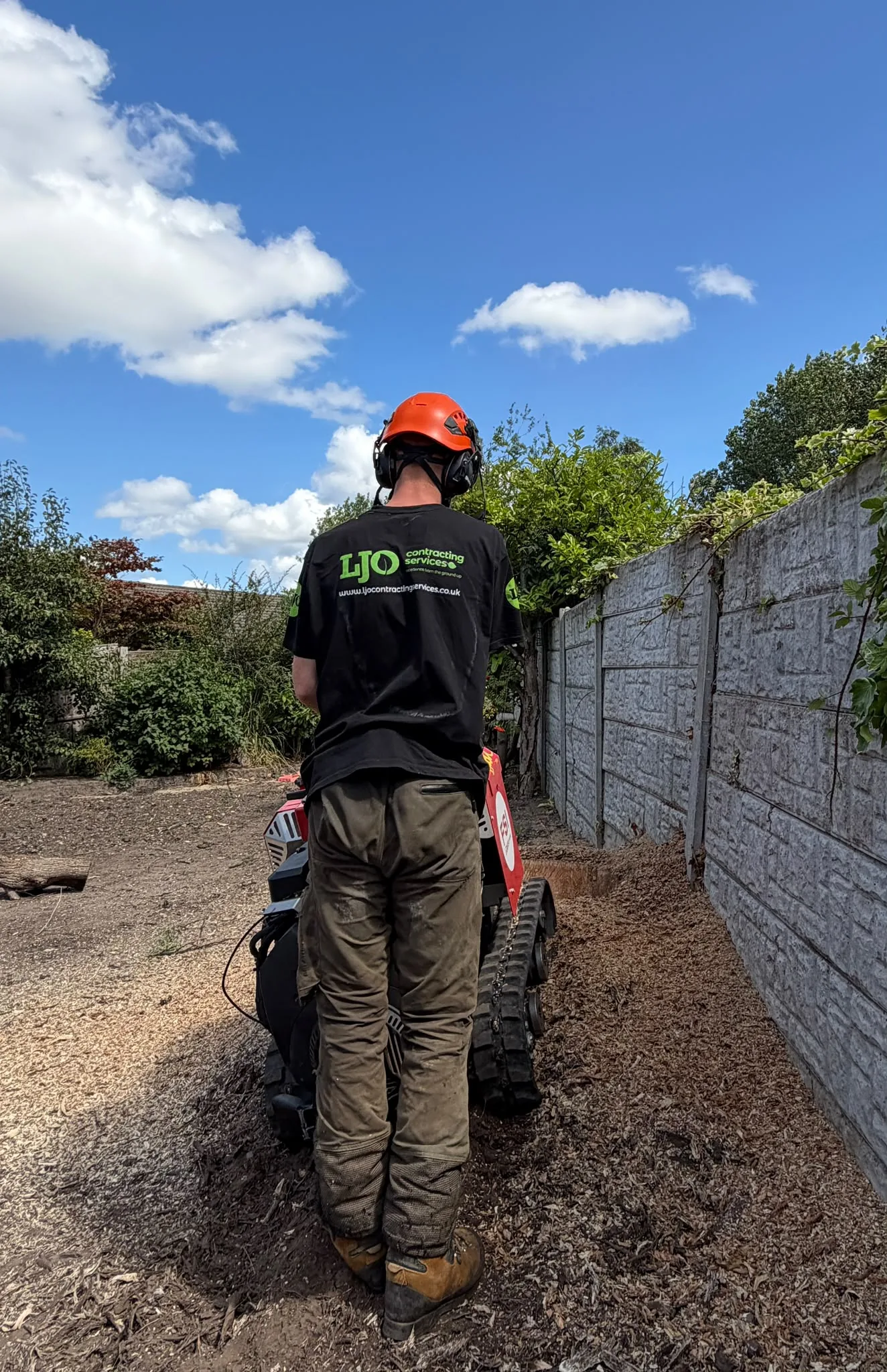 An LJO operative in branded workwear and helmet operating a tracked stump grinder alongside a garden boundary